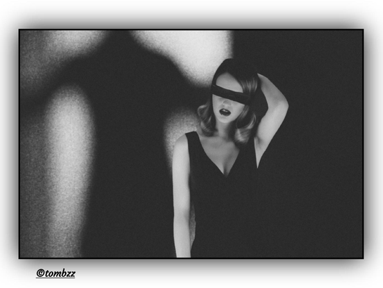 Black-and-white photograph depicting a young woman with her eyes covered by a black blindfold. Her wavy hair appears dark, though the exact color is difficult to determine due to the grayscale. She holds one hand slightly raised as if attempting to untie the blindfold, creating a sense of tension and subtle symbolism. Her lips are prominently highlighted and slightly parted, adding emotional depth to the image. The woman wears a black spaghetti-strap dress and stands against a background split into two contrasting parts: one light, one dark. In the light section, a looming, shadowy figure emerges — its head unseen, presence unknown, yet overwhelming — standing behind the woman and intensifying the mood of unease and ambiguity.