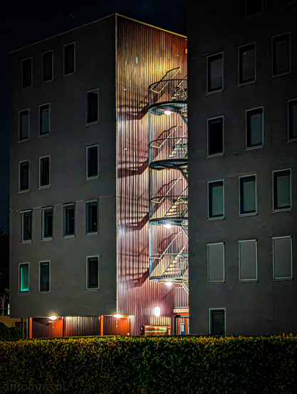 The exterior of two residential buildings separated by a central illuminated section. The dominant colors are muted grays of the building facades, contrasting with the warm, reddish-brown and brightly lit central panel. A metallic, open-tread fire escape with multiple landings and a spiral staircase component is attached to this wall. The strong artificial light emanating from unseen sources above and below illuminates the fire escape and casts dramatic, elongated shadows of its structure onto the wooden paneling behind it. 