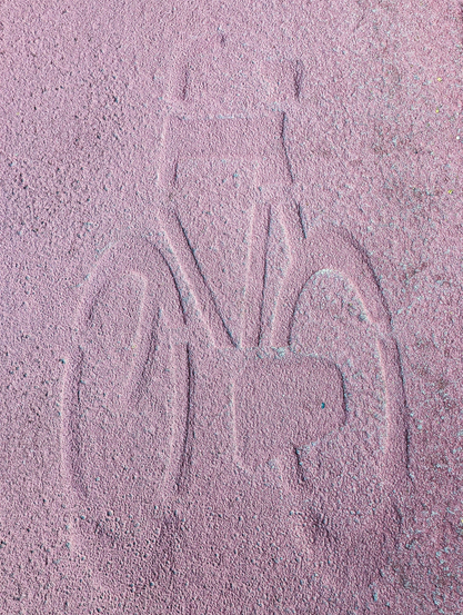 A high-angle shot of a bicycle symbol impressed into a coarse, reddish-pink asphalt surface. All the key components of a bicycle are depicted: two wheels, a frame, handlebars, a seat, and pedals. The lighting creates subtle shadows that give the impression of depth and relief to the symbol. The overall color palette is dominated by various shades of pink and reddish-brown.