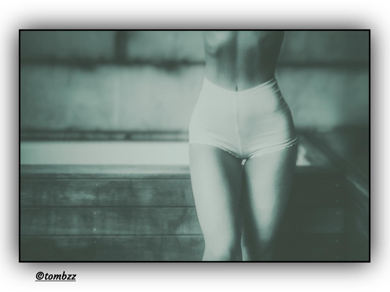 A black-and-white, softly blurred photo of a woman in high-waisted underwear, captured in an artistic close-up that focuses on her hips, thighs, and waist. The subtle, sensual framing set against a raw wooden backdrop creates a contrast between feminine light and its surroundings—a symbol of femininity, delicacy, and strength all at once.