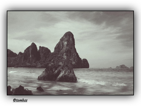A black-and-white coastal photograph. In the foreground, partially submerged rocks are gently touched by waves. In the distance, large rock formations rise from the sea, covered in vegetation, creating a dramatic and picturesque landscape. Above, a lightly clouded sky stretches out. The composition radiates a sense of tranquility and timelessness.