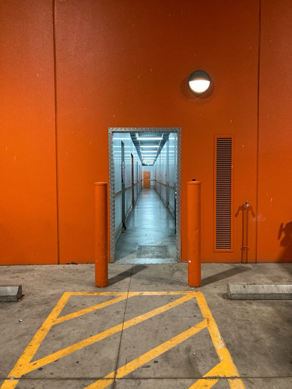 Bright orange wall with a door opening onto a long, brightly lit hallway. There is an  orange door at the distant end of the corridor.