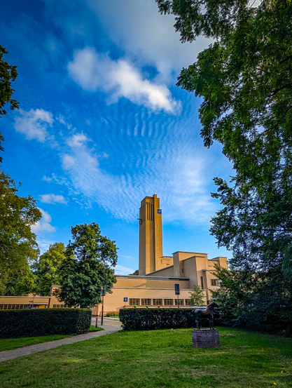 A prominent, tall, light-brown building with a distinctive tower, set against a vibrant blue sky dotted with white clouds. The building has a modern, early 20th-century design, characterized by clean lines and geometric shapes. A blue clock is visible near the top of the tower. 

In the foreground, a well-maintained grassy area leads up to a paved path. Lush green trees frame the top and sides of the image, creating a natural border and adding depth. There are also manicured hedges and other greenery around the base of the building. Many of the clouds exhibit a clear, somewhat parallel, rippled or wavy pattern. 