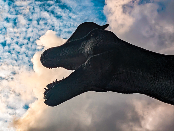 A low-angle shot of a dinosaur silhouette against a vibrant, cloudy sky. The dinosaur, likely a large theropod given its head shape and visible teeth, is entirely in shadow, appearing as a dark, imposing form. Its mouth is open, suggesting a roar or a predatory stance. The sky is a prominent feature, filled with a mix of blue and white clouds. 