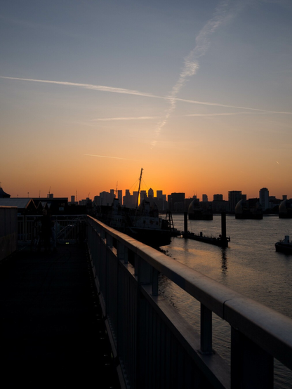 Photo of a distant city skyline silhouetted against an orange-grey-blue sunset sky. In the foreground, a dark railing slides off towards the horizon, reflecting the colours above; on the right, wind-ruffled river water that passes through the shadowed mounds of the Thames Barrier in the middle ground.