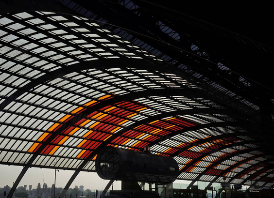A photograph from within a train station, looking up into an overhead canopy structure with a geometric grid pattern of rectangular panels, with some sections showing orange and red coloring while others are clear. A waterfront area with city skyline is visible in the background.