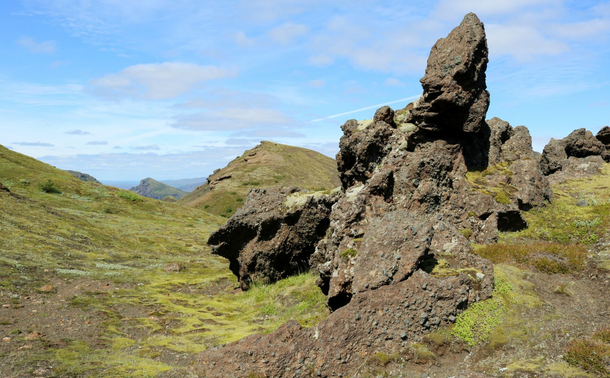 Photograph of a rock formation, light brown with green moss and grey lichen growing in some places, resembling the silhouette of a somewhat deformed creature, with petrified head, arms and body. It was another troll surprised by the sun, according to our Icelandic friend. It sits on top on a mountain, with the ground covered by lime green moss, lichen and alpine plants, facing other mountains in the distance, under a very blue sky.