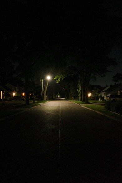 Color photograph looking down a straight residential street at night. The foreground and nearest part of the street is dark, and a ways down a streetlight illuminates a patch of street and some lawns and trees, beyond which it'd dark and then there's another streetlight, seemingly repeated into the distance as far as you can see