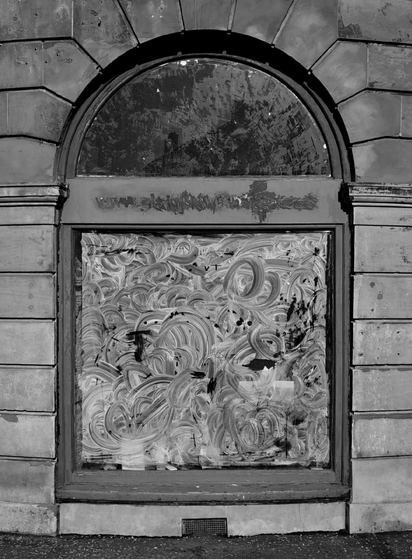 A black and white photograph of an arched storefront window. The glass is covered from the inside in swirling paint patterns to block the view. Some name or text above the window has been scraped off. The building wall shows large weathered stone blocks.