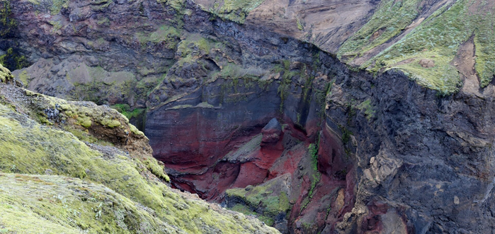 Photograph of a colourful canyon wall seen from the top on the opposite side. The vertical rock is black, grey, brown and vivid red, with patches of green moss. The ground where the photographer is standing is visible on bottom left and is entirely covered by a carpet of light green moss.