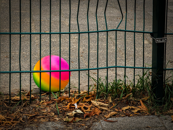 A colorful ball seen through a dark green metal fence. The ball, which is bright pink, orange, and yellow, is resting on a patch of ground covered with dry, brown leaves and some green grass. Behind the fence and the ball, there's a light-colored asphalt playground. The fence itself is made of vertical bars with a horizontal bar across the middle, and it appears slightly bent in places. To the right, part of a thicker, dark post of the fence is visible with a metal clasp or lock mechanism. 