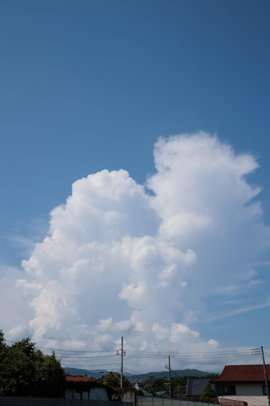 Fluffy white clouds tower in a bright blue sky, above rooftops and power lines, with distant mountains in the background.