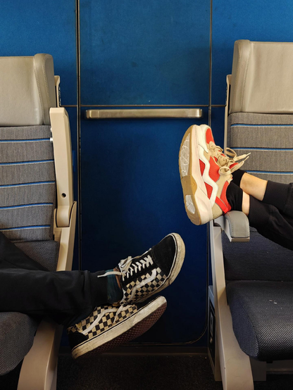 Two people sitting across from each other on a train with their feet up, wearing sneakers - one person in checkered black and white Vans-style shoes, the other in white and red athletic sneakers. The train interior has deep blue walls and gray patterned seats.