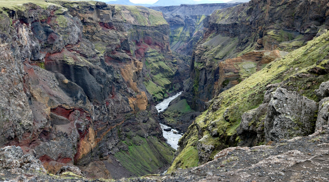 Photograph of a colourful canyon, taken from a standpoint in a detour, where the canyon is bending, allowing to have a view almost as if looking into a corridor. The vertical walls of the canyon are grey, brown, orange, red, all in different hues, and patches of green and lime green moss. The river at the bottom looks silver, reflecting the white cloudy sky. As often with canyons, it doesn't look real.