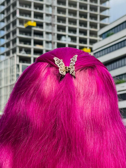 A view of a person's back with vibrant pink hair, styled with a gold butterfly hair clip. In the background, there's a construction site with tall buildings.