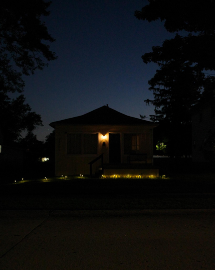 Color photograph of a small house on a dark street at night. The house and trees on either side of it are silhouetted against a deep blue and pink dusky sky, with a porch light and some solar lights on the lawn breaking up the darkness in the foreground