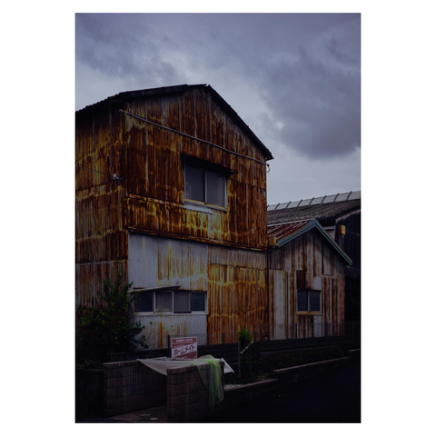 This color photo captures an aging, rust-covered corrugated metal building under a cloudy, foreboding sky—likely in the calm before a typhoon. The building has a weathered, two-story structure, with streaks of rust giving it a fiery appearance against the gloomy blue-grey sky. In the foreground is a trash area partially covered by a green mesh net, draped haphazardly like it's already surrendered to the wind.