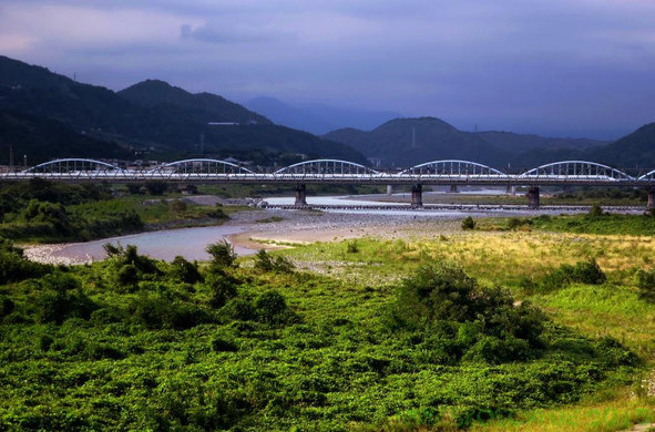 Ein Shinkansen Hochgeschwindigkeitszug passiert auf seiner exklusiven Trasse eine Brücke über den Fuji River bei Fuji City.
Diese Brücke wird gerne als Fotoperspektive mit dem berühmten Vulkan dahinter genutzt. 
Leider verborg sich dieser schüchtern hinter Wolken. 
Die Brücke ist aus der Ferne vor einer Bergkette zu sehen.
Im Vordergrund schlängelt sich ein nicht gefülltes Flußbett durch eine grüne Landschaft.
Der Himmel sieht etwas nach Gewitter aus. 