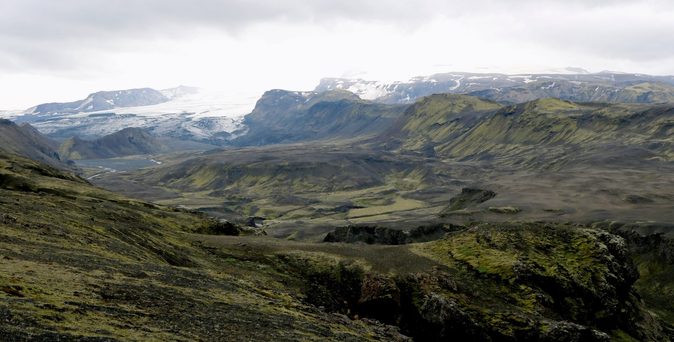 Photograph of a valley of bare brown and green moss covered rock, between high mountains covered with snow and a white glacier, lost in the fog. A river at the bottom of the valley can be seen coming from the glacier on the left. This is an extension of the glacier named Mýrdalsjökull.