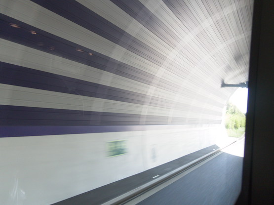 Das Ende eines schwarz-weiß gestreiften Autobahntunnels. Rechts im Bild das Tunnelende mit Schilderbrücke und etwas Grün am Straßenrand sowie der Rahmen vom Busfenster.