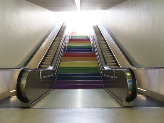 Das Foto zeigt eine Treppe deren Stufen in Regenbogenfarben bemalt sind, rechts und links davon sind Rolltreppen. Die Umgebung ist hell beleuchtet, alle Treppen enden im Licht.
The photo shows a staircase whose steps are painted in rainbow colors, with escalators to the right and left. The surroundings are brightly lit, all the stairs end in light.