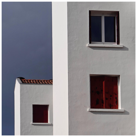 Two white Mediterranean-style buildings against a blue sky. The building on the right and in front has two windows. The building in the background on the left features a clay tile roof and a single window with closed red shutters.