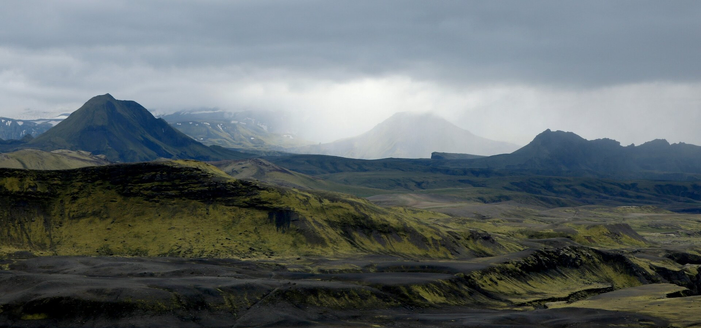 Photograph of a conical mountain lost in the fog under a grey sky but illuminated by a kind of white light coming from the sun. Other mountains around it are darker green and in the shadow. Closer to the photographer in the bottom half of the picture are black hills covered by luminescent green moss.