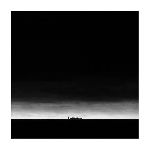 A square-format black-and-white photograph showing a container ship on the distant horizon, centered against a dramatic, dark sky. The image is composed in strong contrast: the sea and sky dominate most of the frame in near-black tones, with a thin horizontal band of lighter sky at the horizon, separating water and clouds. The ship is silhouetted against this narrow bright strip, appearing small and isolated in the vastness of the scene.