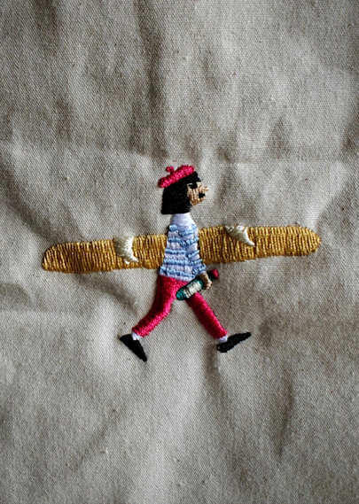 A photograph of a bag, showing a small embroidered patch on light gray fabric depicting a figure with black hair, red baret, blue shirt, red pants, and black shoes. The figure is walking and carries a very large golden baguette.
