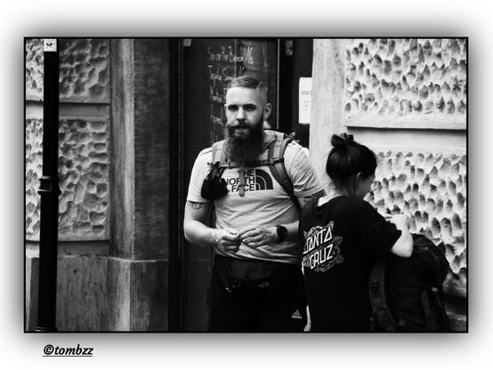 A woman and a man stand in front of a bar entrance, next to a textured concrete wall. The woman is seen from behind—she wears a backpack and a t-shirt with the "Santa Cruz" logo, her hair tied up in a bun or ponytail. The man, also carrying a backpack, looks directly into the camera lens; his gaze is clear, focused, slightly contemplative. He has a short beard and wears a t-shirt with "The North Face" logo. Behind them, there's a chalkboard listing cocktails: "Sea of the Beach," "Tequila Sunrise," "Mojito," and "Frozen Daiquiri." The photo is monochrome, street-style, thoughtfully composed to capture a moment of tension and silence between movement and stillness.