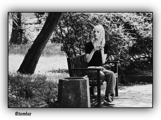 A young woman sits alone on a wooden bench in a park. She's dressed in a black t-shirt, jeans, and black sneakers with white laces. Her hair is loose and falls naturally over her shoulders, with two decorative hair clips placed on either side — more of an accent than a practical hold. Her face tilts slightly downward, eyes focused and thoughtful as she gazes at the smartphone held in both hands, seemingly disconnected from her surroundings. Beside the bench stands a concrete trash bin. In the background are shrubs and the trunks of trees, highlighted by soft light that gently brings out their textures. A black-and-white street portrait that captures a quiet, melancholic moment of everyday life.