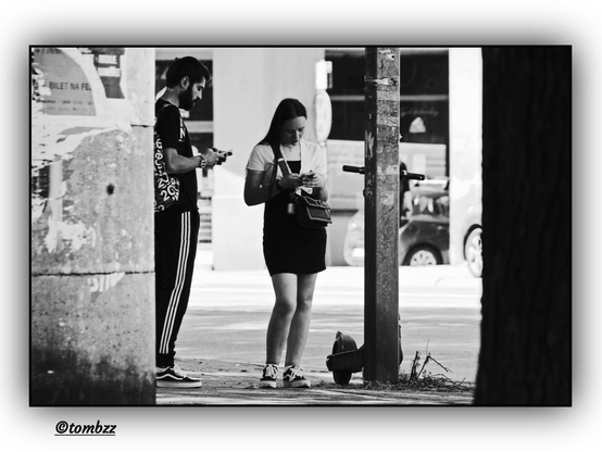 A young woman and a young man stand on a city sidewalk, both absorbed in their smartphones — a snapshot that reflects the rhythm of modern life. The woman wears a short-sleeved top and a skirt, with a boldly designed bag slung over her shoulder; her hair is loose, flowing smoothly down her back. The man sports a patterned shirt and vertically striped pants; his beard is neatly groomed, his posture upright, head tilted slightly toward his phone.
At their feet lies a scooter, casually dropped near a metal pole — perhaps abandoned in haste. The background sets a classic urban scene: building facades, shop windows, a parked car, and a section of a tree trunk visible on the far right of the frame. Captured in black and white with a documentary eye, the image gently reflects the rituals of city life today.
