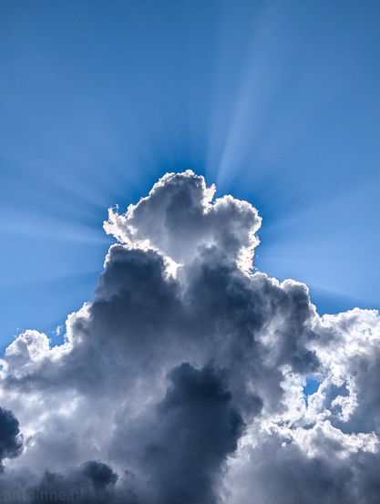 A sky scene dominated by a large, dark, and somewhat ominous cloud formation, occupying the lower two-thirds of the frame. The cloud has various shades of gray, from very dark in the foreground to lighter, more illuminated areas towards the top.

Behind and above this main cloud, a brilliant burst of sunlight emanates, creating striking rays that fan out into the clear blue sky. The sun itself is hidden behind the cloud, making the cloud's silhouette sharply defined against the radiant backdrop.