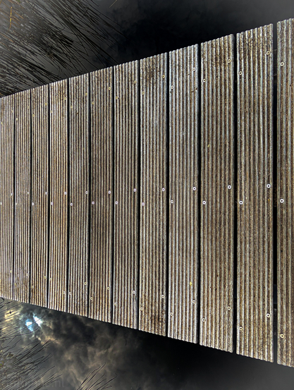 A wooden boardwalk extending over dark, reflective water. The boardwalk is composed of numerous long, narrow wooden planks laid parallel to each other. Each plank has a ridged or grooved surface, and they are secured with visible silver-colored screws or bolts arranged in a line down the center of each plank. To the left side of the boardwalk, the water reflects a bright, light source, creating a shimmering, almost ethereal glow on the dark surface. 