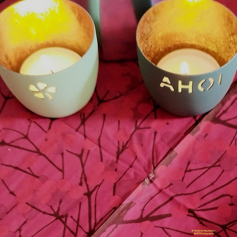 Two turquois-green tealight holders on a red napkin with red, and brown patterns. The left candleholder is of lighter colour, and has a four-leaved clover cut into it, the right one is of darker colour and has the German word 'Ahoi' cut into it. Both tealight holders are golden on the inside.