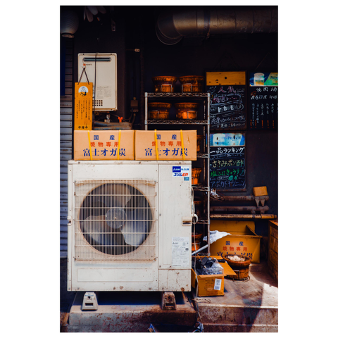 This color photo captures a compact, cluttered corner likely outside a Japanese izakaya or restaurant in Nagoya. At the center is a large, aging outdoor air conditioning unit, slightly stained from years of use. On top of it are three stacked cardboard boxes labeled 富士オガ炭 (Fuji Ogatan charcoal), a type of compressed charcoal used for grilling. Behind the AC unit is a metal shelf holding clay grilling pots and more supplies. To the right, blackboard menus with handwritten chalk lettering list food items, drinks, and lunch sets. The space is dimly lit, but pockets of sunlight highlight the earthy tones of charcoal dust, rust, and ceramic.