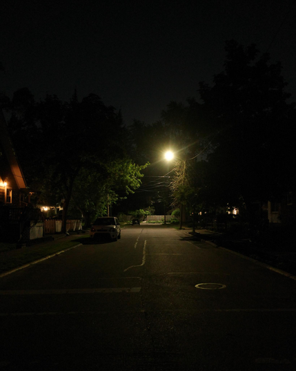 Color photograph looking down a straight residential street at night. A streetlight a little ways down illuminates part of the street and some trees and another light a little further shines on a fence where the street dead-ends. Trees loom up along the side of the street and beyond it, black against a darkly glowing night sky