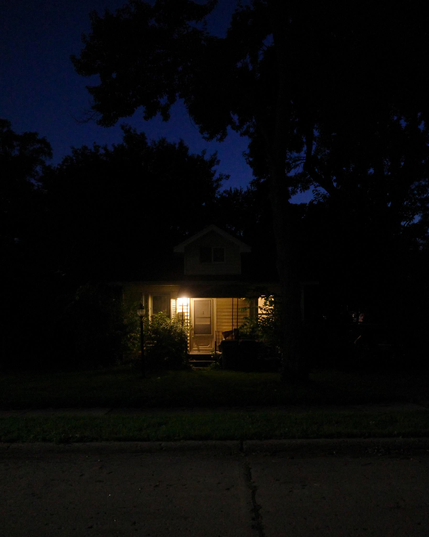 Color photograph looking at the front of a small bungalow at dusk, a porch light warmly illuminating the porch, and the light all around dim enough that the it looks the porch is nestled back into a thick growth of trees and bushes grown up in front of and all around the house, deep blue dusky sky showing through patches in the trees overhead