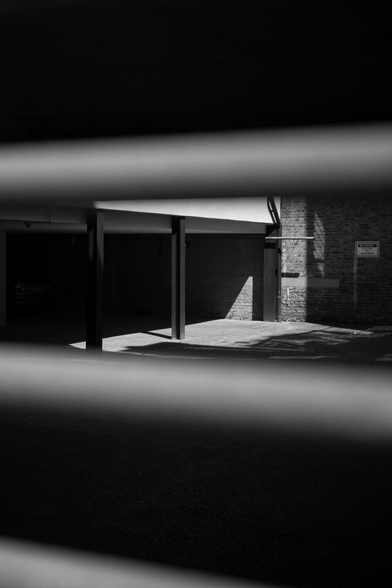 Monochrome photo of what is probably a small enclosed car park, a brick wall at the back, dark metal pillars holding up a white building at the left, sun coming in from somewhere above. This is seen through a slot in a barrier of some kind, out-of-focus in the extreme foreground – indistinct grey stripes across a dark background.