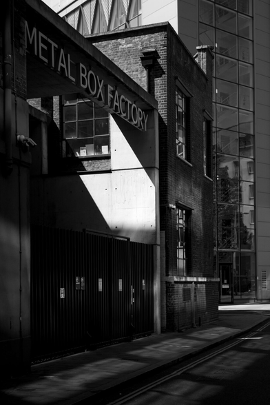 Black and white photo of a two-storey boxy brick building joined onto what is basically a large open concrete box with the words "METAL BOX FACTORY" inside its top edge and a black metal fence and double gate at its base. There's a gap in this structure around a window in its neighbour and slanted sunlight floods through across that far interior wall. Bright light also bounces from elsewhere onto the otherwise shadowed front wall of the brick building, illuminating a patch and reflecting from three more windows. Standed angled in the background is a modern boxy block, rising out of shot, a column of glass panes running up its shaded front and triangular window frames in the sun-blasted wall visible at the top of the frame.