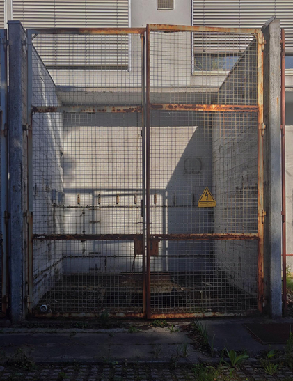 A photograph into a small empty outside area with a metal security gate with wire mesh panels and visible rust, featuring a yellow triangular warning sign about high voltage. The gate has horizontal support bars and is mounted between concrete posts, with some vegetation growing at its base and white concrete visible in the background.