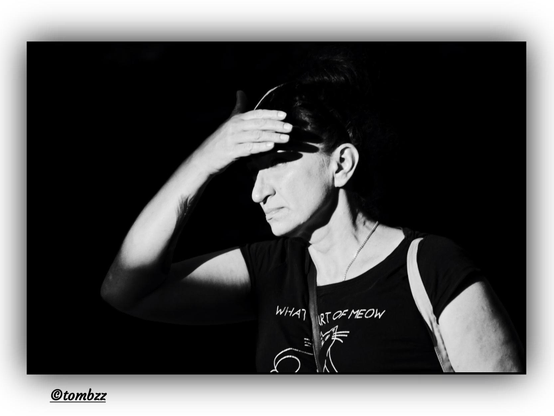 Black and white street portrait depicting a woman standing in the intense light of the setting sun. She is wearing a black t-shirt with a prominent cat print and the text “WHAT PART OF MEOW”. A chain is visible around her neck. Her hand is raised to her forehead, shielding her eyes from the harsh light. Strong contrasts of light and shadow highlight the texture of her clothing, her gesture, and facial features. The dark background emphasizes her silhouette, making her stand out clearly in the composition.
