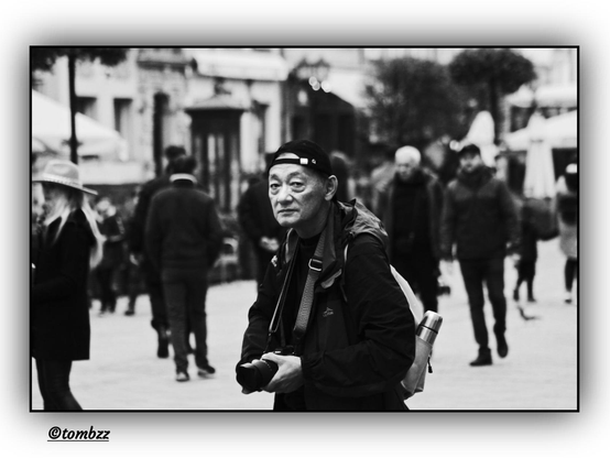 A black-and-white street photograph depicting a vibrant urban scene along a promenade. At the center of the frame stands a photographer — a man wearing a baseball cap turned backward, holding a camera in his hands. His facial expression appears startled, as if he’s just realized he’s become the subject of another photographer’s shot. He carries a backpack with a water bottle or thermos attached, dressed in casual attire suited for walking and shooting outdoors.
The background is full of life: passersby stroll in various directions, some with bags or backpacks. A woman in a hat stands out among the crowd, adding flair to the composition. Behind them, the scene includes building façades, trees, and street umbrellas — a bustling urban space pulsing with contrast and energy. The image emphasizes the interaction between the observer and the observed.