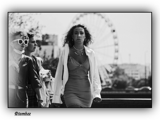 A black-and-white photo shows three girls walking together. The central figure wears a short dress with a white shirt draped over her shoulders and a necklace with beads. Behind her, another girl is holding a plastic cup and has a sweatshirt slung over one arm. The third girl, on the right side of the frame, wears sunglasses, a blouse with a deep neckline, pants, and a necklace with beads. In the background, a large Ferris wheel, a building, parked cars, and typical urban scenery are visible. The left side of the image is slightly overexposed.