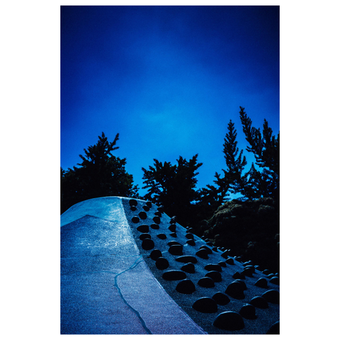 The photo captures an abstract and surreal scene of a playground structure resembling Mount Fuji, located somewhere in Nagoya. The concrete surface is textured and painted in cool blues, with a jagged black line running like a fault crack down its middle. Rows of black, dome-shaped climbing grips line the slope. Tall pine trees form a dark silhouette against a deep cobalt-blue sky, giving the whole scene an uncanny, almost otherworldly feel.