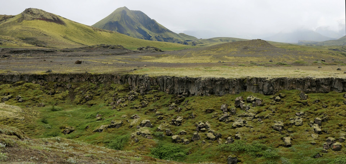 Photograph of green hills and brown plain at the top half of the picture, suddenly cut vertically in the middle, forming a brown rocky wall a couple of meters high. In the bottom half, small rocks have fallen from the wall on a greener terrain, with moss and green plants and bushes are growing.