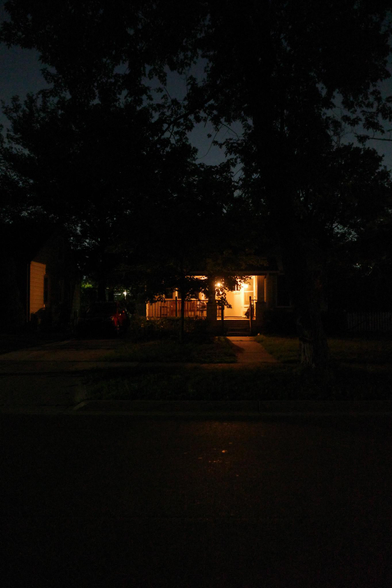 Color photograph looking across a darkened residential street at night at the front of a small house whose covered porch is lit brightly by golden light, some of the light dimly spilling out over the sidewalk and onto the street, with trees looming all around silhouetted against a brightly glowing night sky