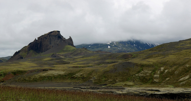 Photograph of a brown small mountain with a strange shape, as if a bite was taken off the top, looking more or less like a rhinoceros head according to most hikers in the group. Although one hiker mentioned a dung beetle, the one with a horn... But our guide insisted the mountain was named Einhyrningur, meaning "the unicorn". The mountain is in the middle of brown and green hills. Behind, a bigger green mountain with white snow is almost all hidden behind fog and clouds. At the bottom is green and brown grass.
