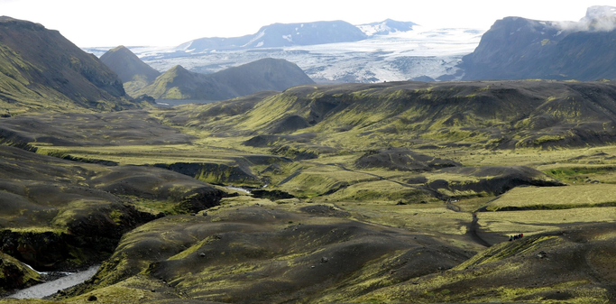 Photograph of a valley of bare brown rock covered with green moss, between high mountains with snow and a white glacier, at the top of the picture. This is an extension of the glacier named Mýrdalsjökull. A river is running at the bottom of the valley, forming a small canyon, and a trail is crossing the entire landscape, with groups of hikers on it.