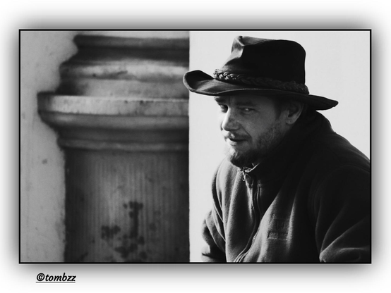 Black and white photograph featuring a man wearing a fedora or western-style hat, seated on a low wall next to a large cylindrical column with a textured surface. Dressed in a fleece jacket, he gazes toward the photographer — his expression focused, contemplative, perhaps slightly melancholic — lending the scene a reflective tone. His beard adds distinctiveness and character to his face. In the background, part of a building facade creates a typical urban setting. The image evokes a street photography vibe, with a classic, almost cinematic atmosphere.